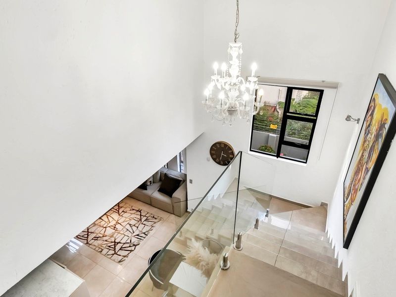 A bright, double-volume entrance hallway featuring a modern glass balustrade, tiled staircase, and an elegant chandelier, overlooking the living area below.