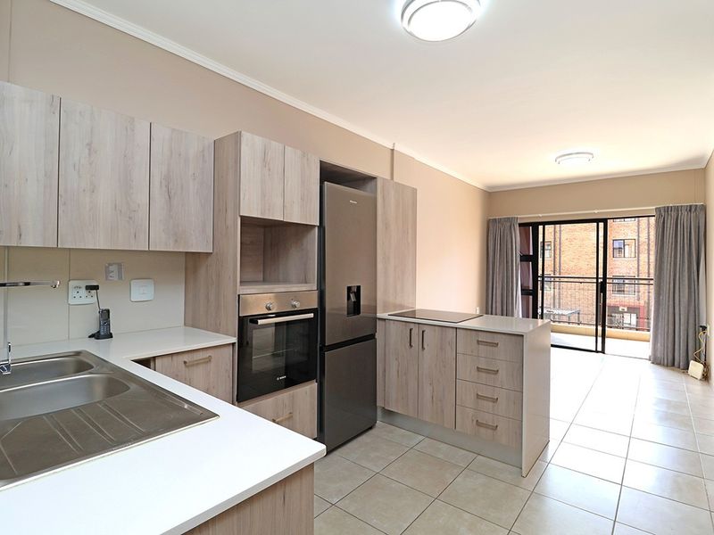 Modern open-plan kitchen featuring light wood-grain cabinetry, built-in oven, cooktop, and tiled flooring, leading to a balcony with sliding glass doors.
