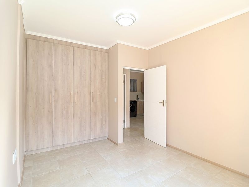 A bright, empty bedroom featuring neutral tiled flooring, built-in wooden cupboards, and a view through to an adjacent utility area.