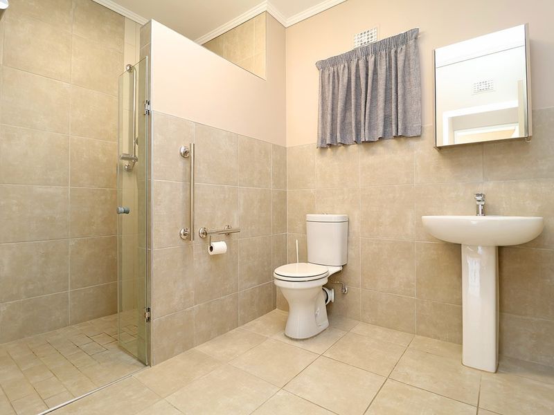 A clean, functional bathroom featuring a walk-in shower with glass screen, pedestal basin, and safety grab rails, finished with neutral ceramic tiling.