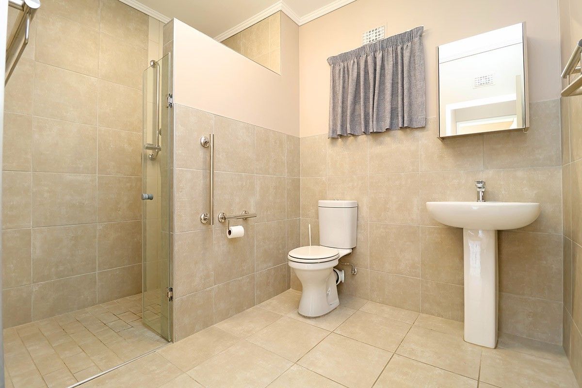 A clean, functional bathroom featuring a walk-in shower with glass screen, pedestal basin, and safety grab rails, finished with neutral ceramic tiling.
