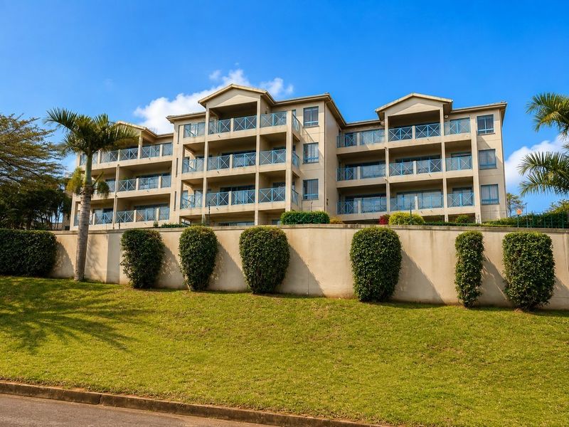 Rear view of a multi-story apartment complex featuring private balconies with glass balustrades, set above a well-maintained retaining wall and landscaped garden area.