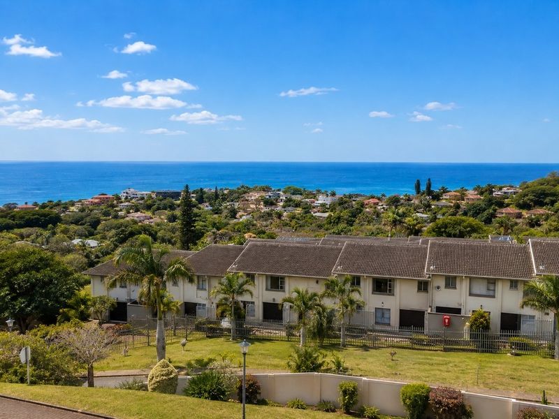 An elevated aerial view of a residential apartment complex featuring tiled roofs, manicured lawns, and perimeter fencing with a scenic ocean backdrop.