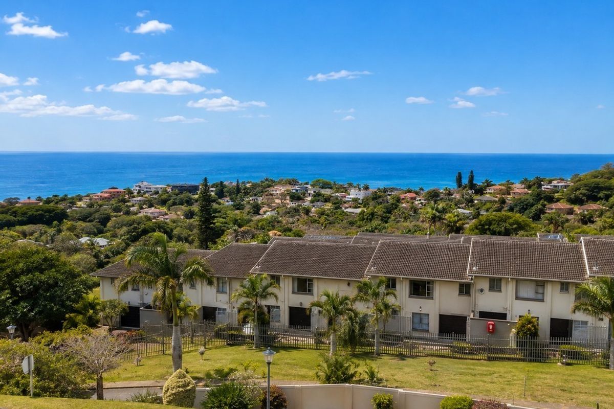 An elevated aerial view of a residential apartment complex featuring tiled roofs, manicured lawns, and perimeter fencing with a scenic ocean backdrop.