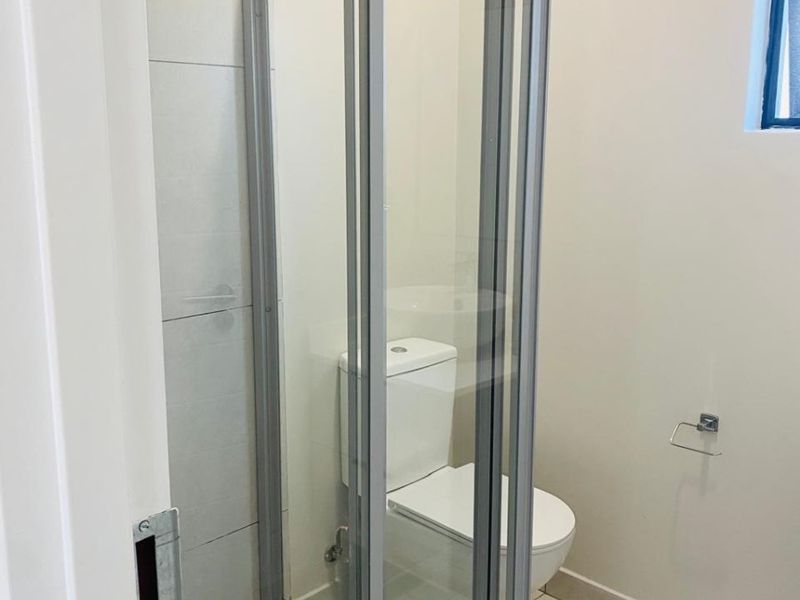 A compact bathroom featuring a glass-enclosed shower with a rain shower head and a standard toilet, finished with neutral floor and wall tiling.