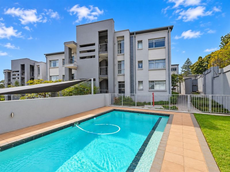 A well-maintained communal swimming pool area featuring a tiled deck, shade sail, and secure perimeter fencing within an apartment complex.