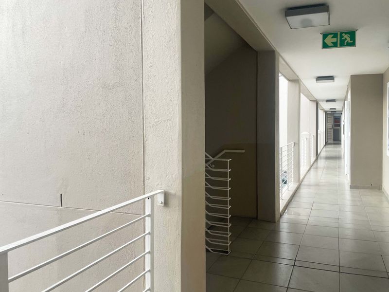 A clean, well-lit apartment building hallway featuring tiled flooring, metal safety balustrades, and clear emergency exit signage.