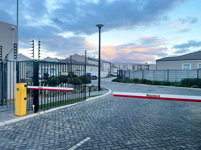 Secure residential complex entrance featuring a paved driveway, automated boom gates for residents and visitors, and perimeter electric fencing.