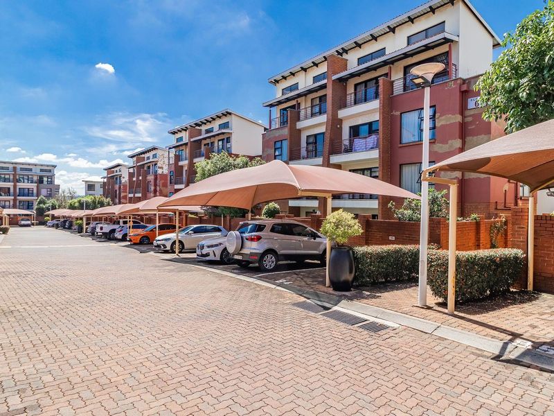 Exterior view of a townhouse complex featuring paved driveway, covered parking bays, and multi-story residential buildings with brick boundary walls.
