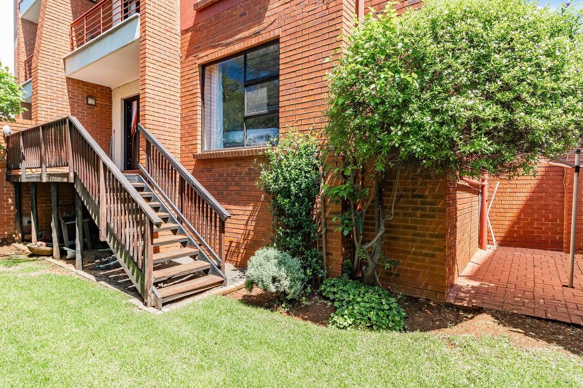 Rear view of the townhouse featuring a brick facade, a raised wooden staircase leading to a balcony, a paved patio area, and a private garden.
