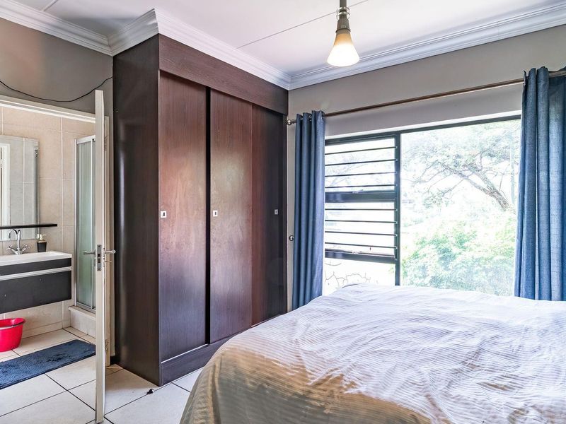 A well-lit bedroom featuring built-in dark wood cupboards, tiled flooring, and an adjoining en-suite bathroom with a modern vanity and shower.