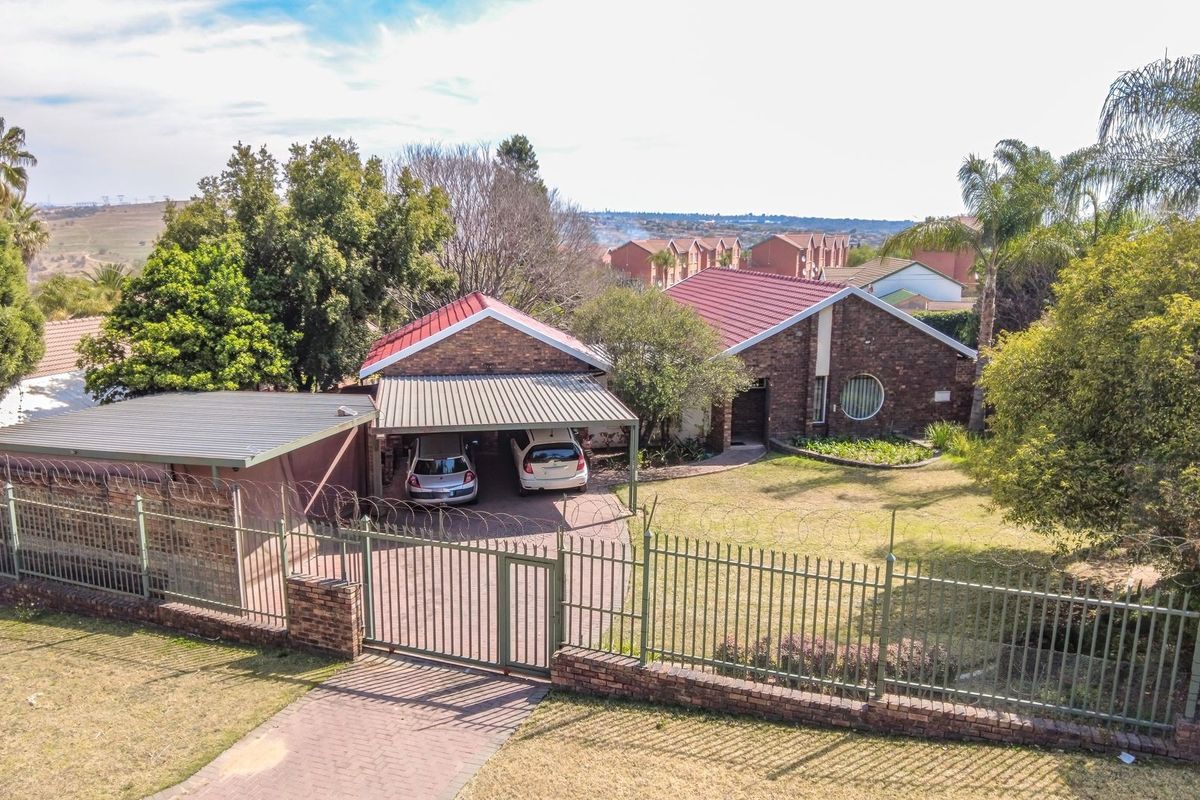 Aerial view of a brick house featuring a metal roof, carport, paved driveway, and secure perimeter fencing with razor wire.