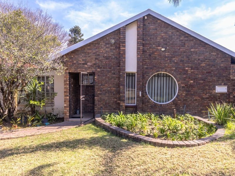 Front elevation of a face brick house featuring a distinctive circular window, security entrance gate, and a landscaped front garden with a paved walkway.