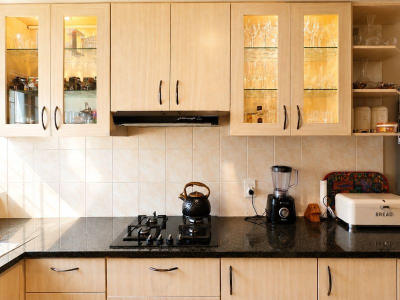 A well-maintained kitchen featuring light wood-finish cabinetry, dark granite countertops, a gas hob, and integrated glass-front display cabinets with internal lighting.