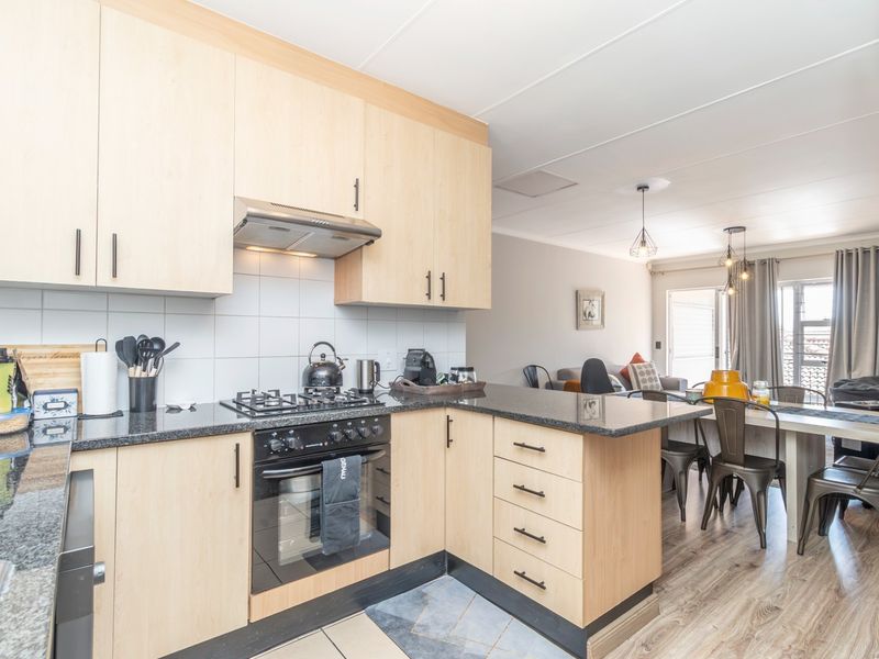 Open-plan kitchen and dining area featuring light wood cabinetry, granite countertops, integrated appliances, and laminate flooring leading to a balcony.