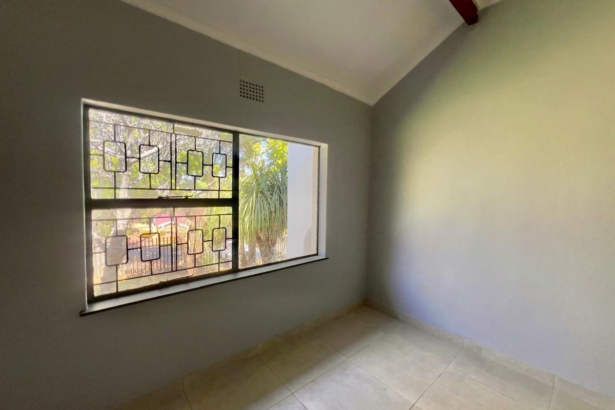 A bright, empty bedroom featuring tiled flooring, a vaulted ceiling with an exposed beam, and a window fitted with security bars.