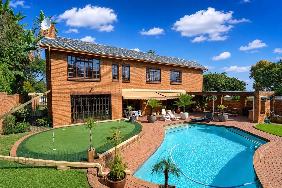 Rear view of a double-story brick house featuring a swimming pool, artificial putting green, and a covered patio area with a retractable awning.