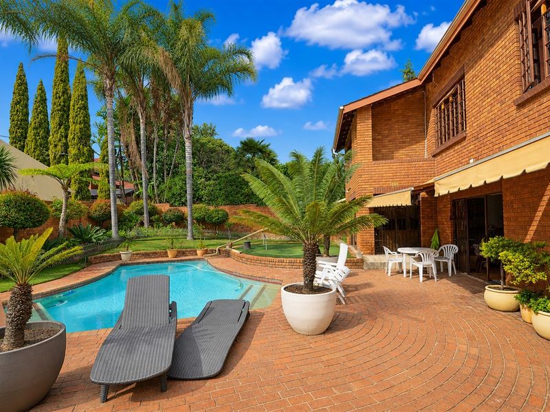A well-maintained outdoor pool area featuring brick paving, a swimming pool, and a brick facade with a retractable awning.