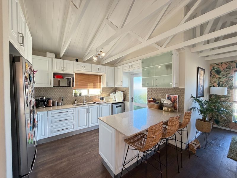 Modern kitchen featuring white shaker cabinetry, stone countertops, a central island with breakfast bar seating, and distinctive exposed timber ceiling beams.