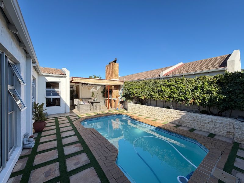 Outdoor pool area featuring a paved surround, stone feature wall, and a covered patio space with a built-in brick braai.