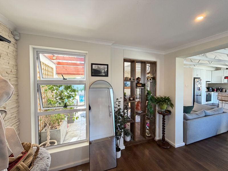 Open plan living area featuring timber flooring, recessed lighting, and a custom built-in timber shelving unit, with views through to the kitchen and patio.