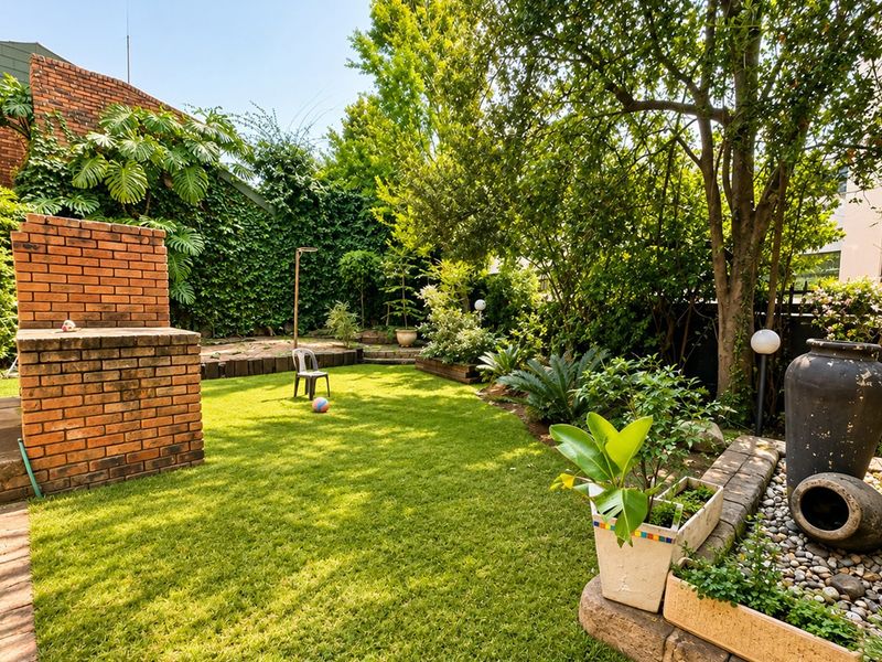 A private rear garden featuring a brick built-in braai, lush lawn, mature trees, and a decorative stone water feature.