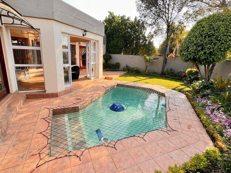 A well-maintained outdoor pool area featuring a tiled patio, swimming pool with safety net, and landscaped garden borders.