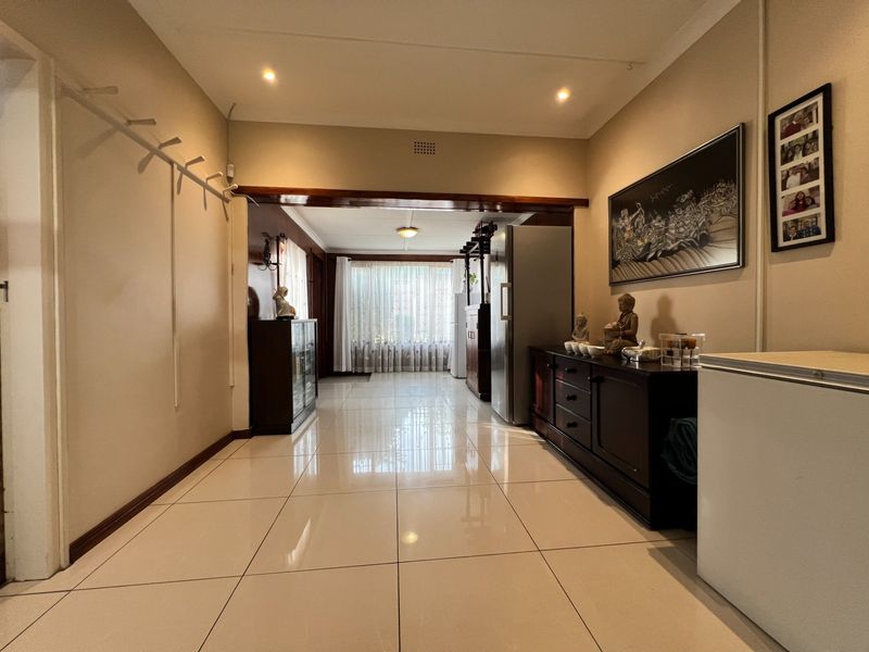 A spacious, well-lit hallway featuring polished light-colored floor tiles and recessed downlighting, leading through a timber-framed opening into an adjacent living area.