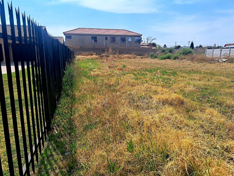 A vacant residential plot featuring a metal spike boundary fence and perimeter wall, with natural grass and vegetation cover.