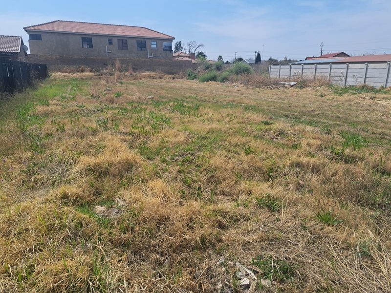 A vacant residential plot featuring dry, overgrown grass and enclosed by precast concrete boundary walls and perimeter fencing.