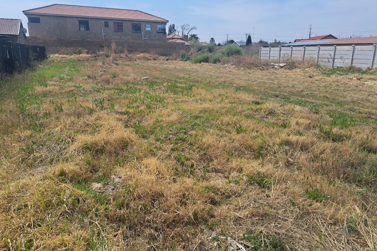 A vacant residential plot featuring dry, overgrown grass and enclosed by precast concrete boundary walls and perimeter fencing.