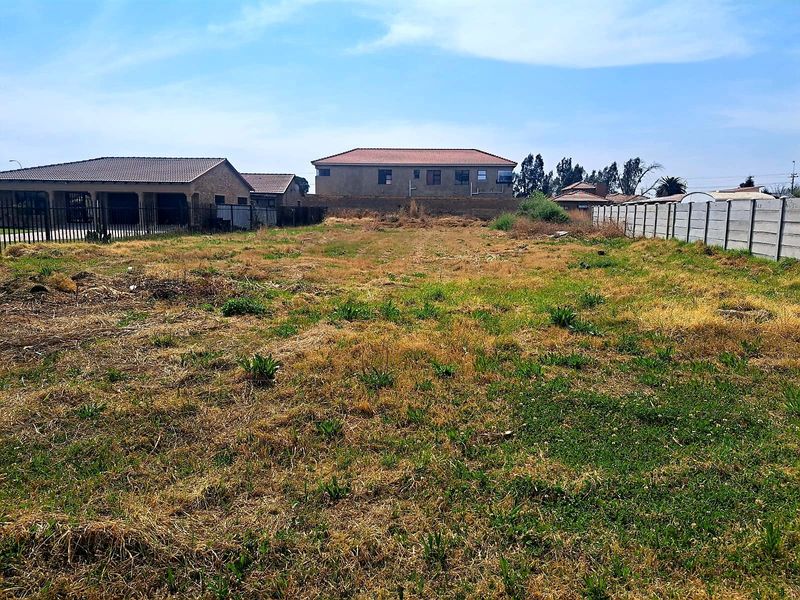 A vacant residential plot featuring natural grass cover, enclosed by a precast concrete boundary wall and metal perimeter fencing.