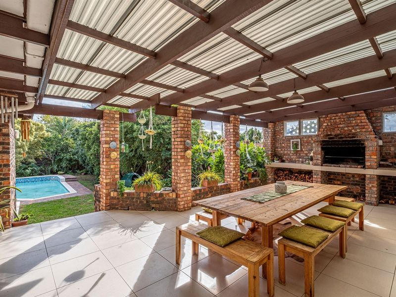 Spacious covered patio featuring a built-in brick braai, timber pergola roof, and tiled flooring, overlooking the garden and swimming pool area.