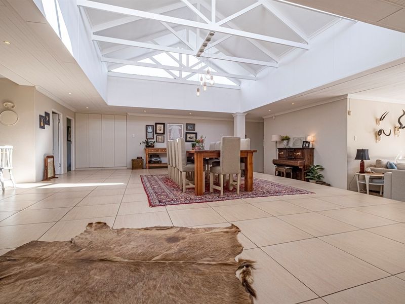 Spacious open-plan living and dining area featuring a high vaulted ceiling with exposed white trusses, recessed lighting, and large format floor tiles.