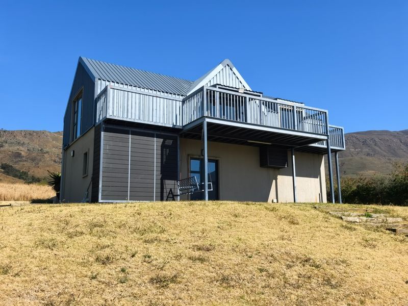 Rear view of a modern two-story house featuring a large elevated balcony with metal railings and a metal roof, set against a mountainous landscape.