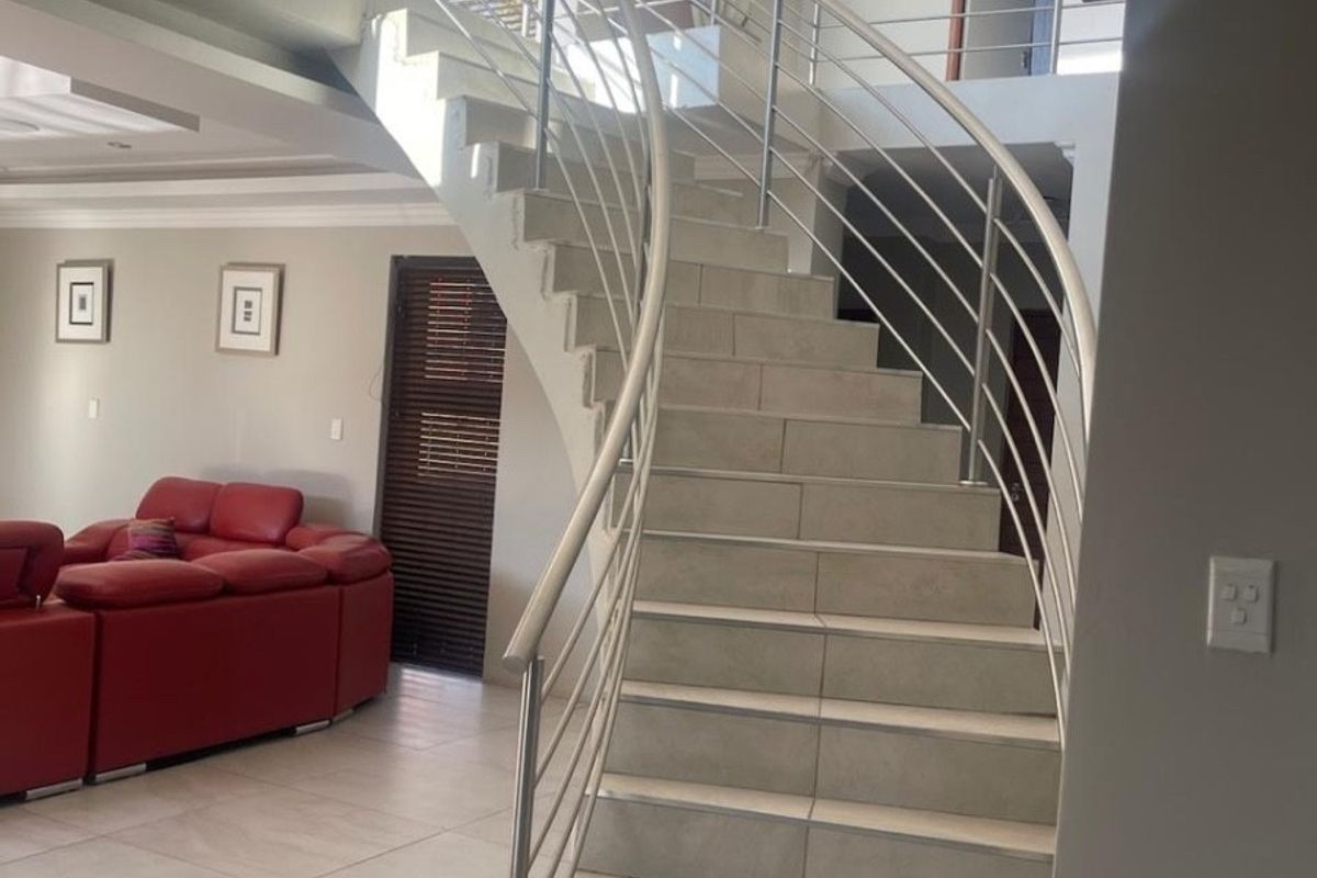 Spacious living area featuring a prominent curved staircase with stainless steel balustrades, tiled flooring, and double volume ceilings.