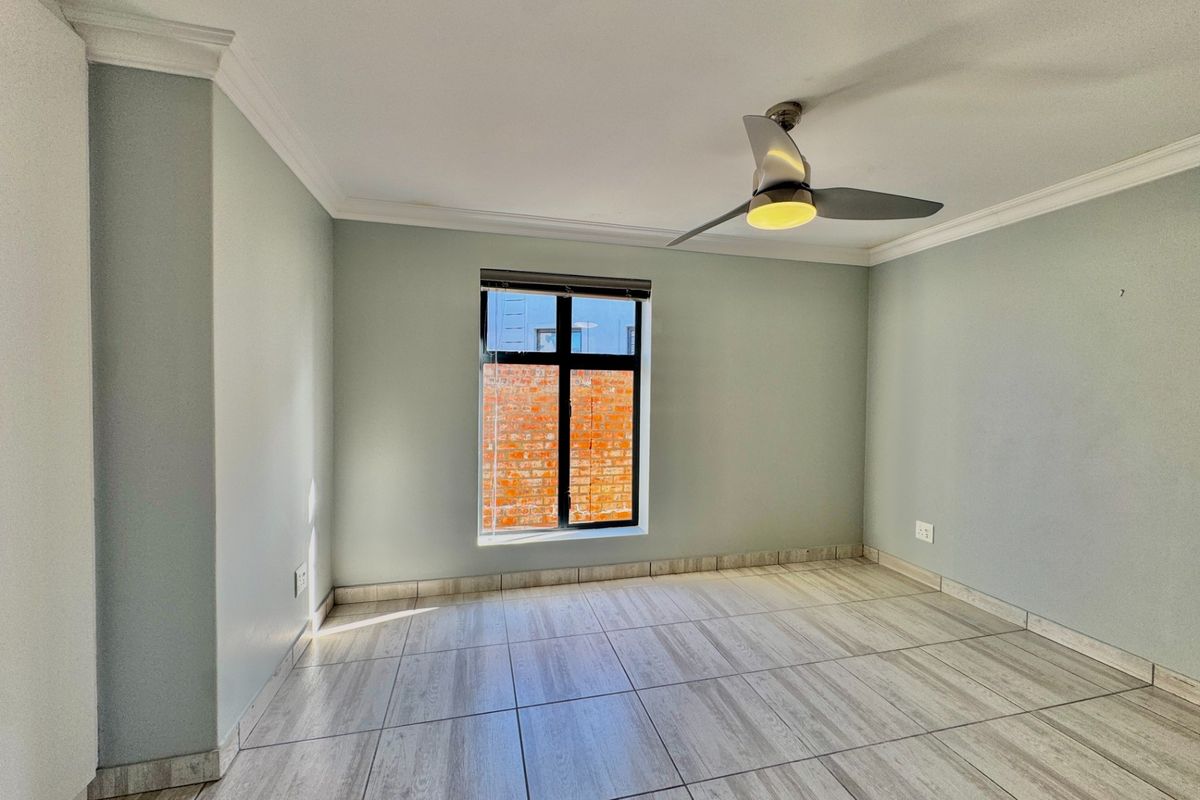 A bright, empty bedroom featuring neutral wall paint, modern wood-look tiled flooring, a ceiling fan, and a window with a view of a brick wall.