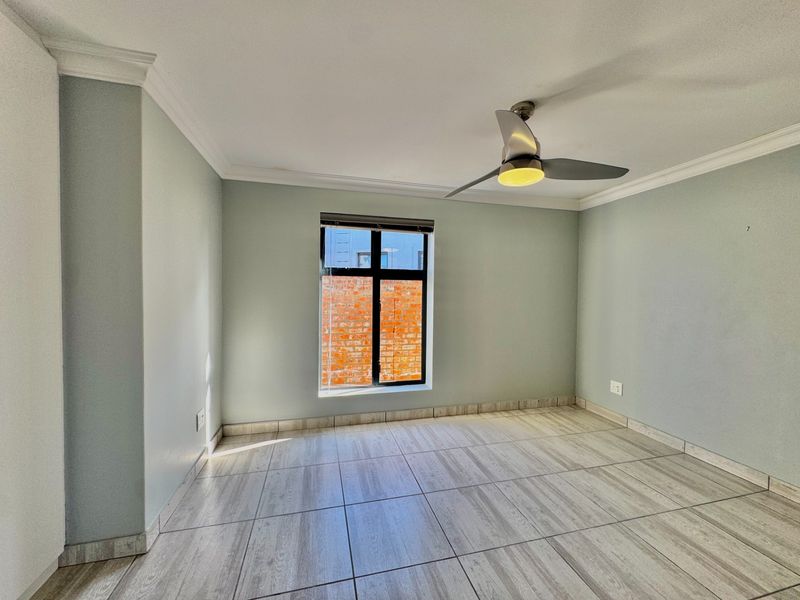A bright, empty bedroom featuring neutral wall paint, modern wood-look tiled flooring, a ceiling fan, and a window with a view of a brick wall.
