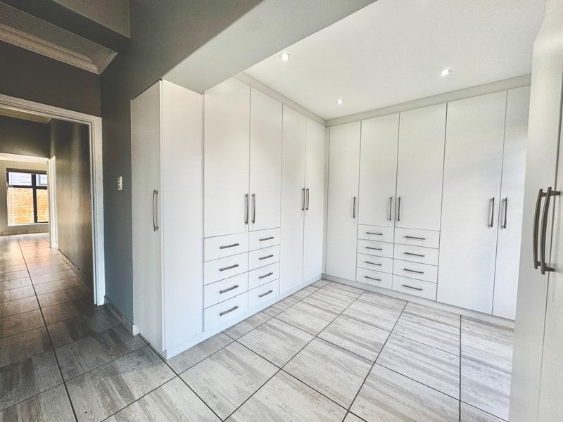 A spacious bedroom featuring extensive white built-in cupboards with modern hardware, recessed lighting, and neutral tiled flooring.