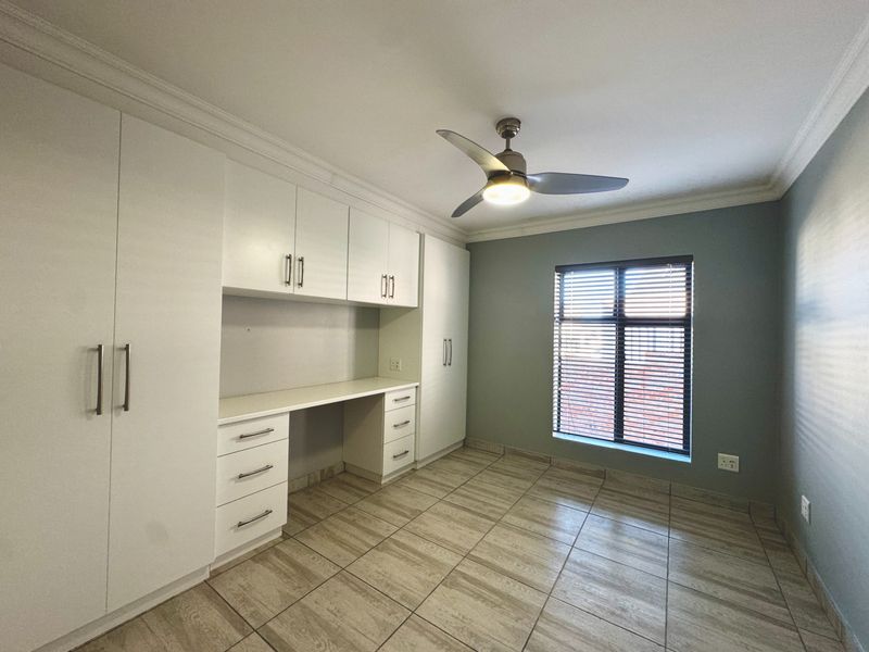 A clean, empty bedroom featuring built-in cabinetry with an integrated desk, tiled flooring, a ceiling fan, and large windows with venetian blinds.