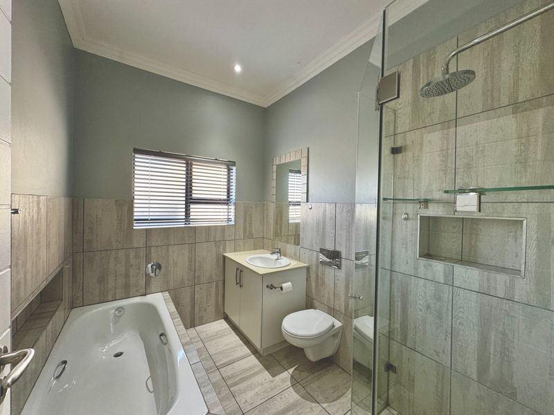 Modern bathroom featuring a built-in bathtub, a glass-enclosed shower with a recessed niche, a wall-mounted toilet, and a vanity cabinet with neutral tiling.