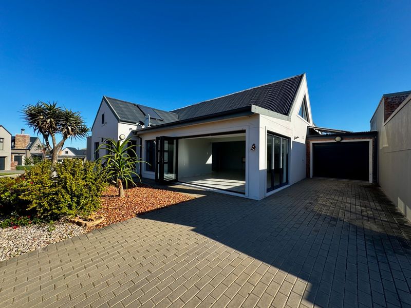 Modern single-story residence featuring a corrugated metal roof, paved driveway, integrated garage, and large bi-fold doors opening to the front garden area.
