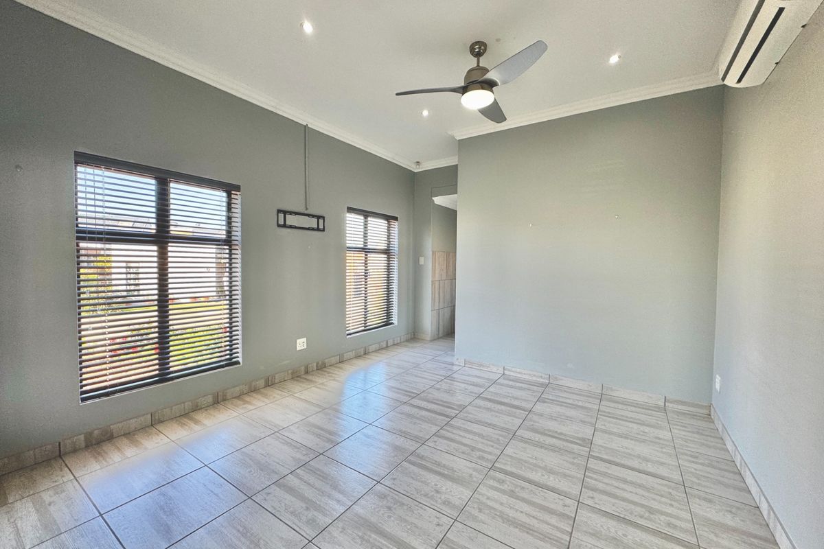A bright, empty living room featuring neutral grey walls, tiled flooring, a ceiling fan, and a wall-mounted air conditioning unit.