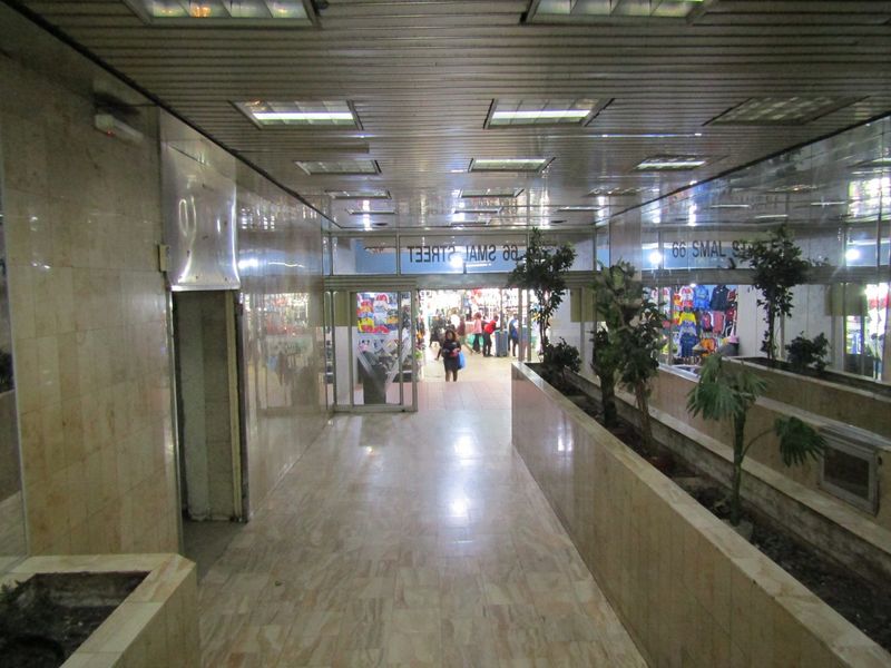 A spacious commercial hallway featuring marble wall and floor finishes, recessed ceiling lighting, and integrated planter boxes leading to a street-level entrance.