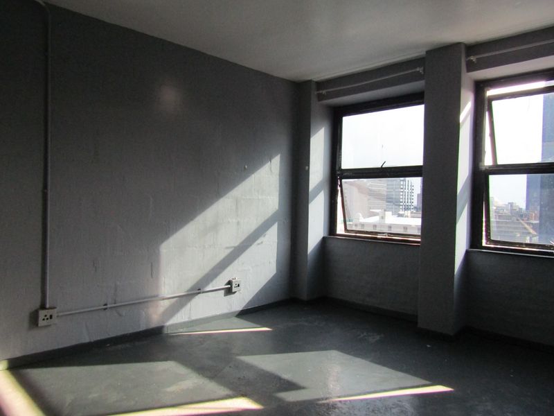 An empty bedroom featuring painted concrete floors, exposed electrical conduit, and large casement windows with urban views.