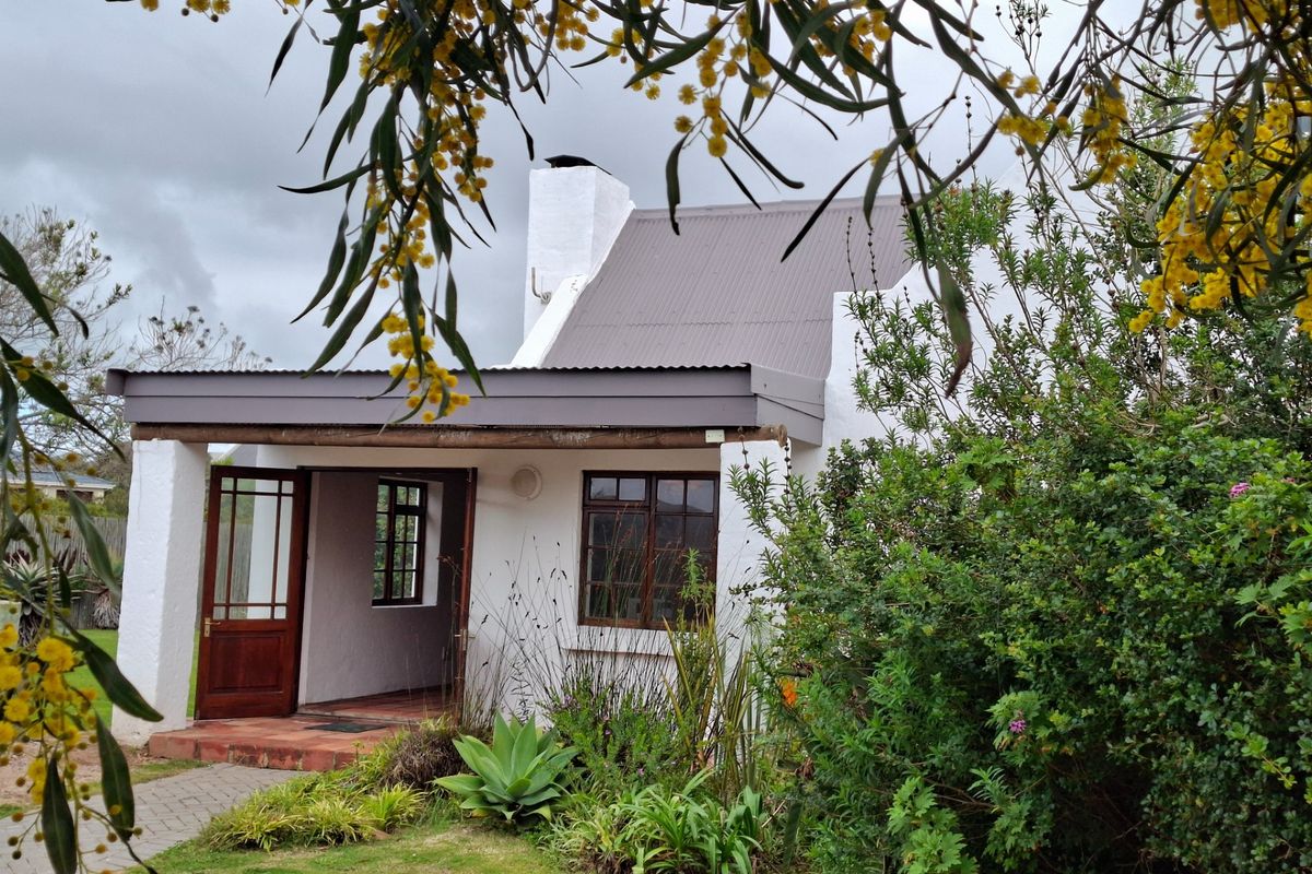 A charming cottage-style exterior featuring a covered patio, corrugated metal roof, and established garden surroundings.