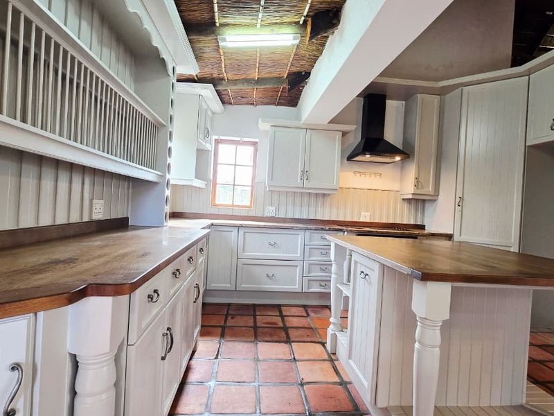 A rustic kitchen featuring white cabinetry, wooden countertops, terracotta flooring, and a unique exposed thatch ceiling with an integrated extractor fan.