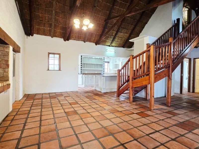 Spacious open-plan living area featuring exposed timber rafters, a thatched ceiling, terracotta flooring, and a prominent built-in brick fireplace with a wooden staircase.