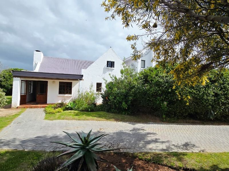 Front view of a charming cottage-style house featuring a corrugated metal roof, covered entrance porch, and a paved driveway set within a garden.