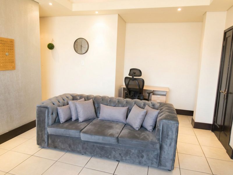 A well-lit living area featuring neutral tiled flooring, recessed ceiling lighting, and clean wall finishes, providing a versatile space for relaxation or home office use.